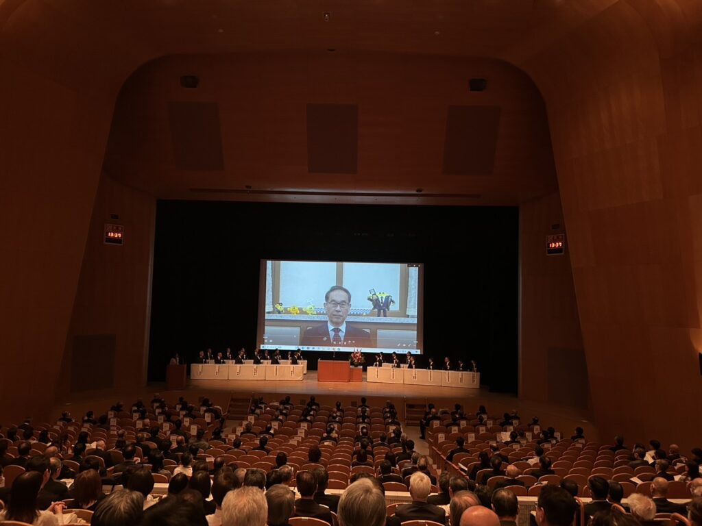 大会会場でスクリーンに大野元裕 第62代埼玉県知事のビデオメッセージが映し出され、参加者が着席して視聴している様子。