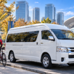 秋晴れのさいたま新都心で、白い大型ジャンボタクシー(実在の「トヨタ・ハイエース ワイド スーパーロング」の貸切タクシー)が歩道脇に停まり、開いたリアゲートの荷室に前輪を外したロードバイク2〜3台が丁寧に積まれている写真。