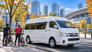 秋晴れのさいたま新都心で、白い大型ジャンボタクシー（実在の「トヨタ・ハイエース ワイド スーパーロング」の貸切タクシー）が歩道脇に停まり、開いたリアゲートの荷室に前輪を外したロードバイク2〜3台が丁寧に積まれている写真。