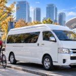 秋晴れのさいたま新都心で、白い大型ジャンボタクシー(実在の「トヨタ・ハイエース ワイド スーパーロング」の貸切タクシー)が歩道脇に停まり、開いたリアゲートの荷室に前輪を外したロードバイク2〜3台が丁寧に積まれている写真。