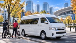 秋晴れのさいたま新都心で、白い大型ジャンボタクシー（実在の「トヨタ・ハイエース ワイド スーパーロング」の貸切タクシー）が歩道脇に停まり、開いたリアゲートの荷室に前輪を外したロードバイク2〜3台が丁寧に積まれている写真。